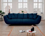 Dunkelblaues, großzügig gepolstertes 3-Sitzer-Sofa mit mehreren Kissen und schrägen Metallfüßen im hellen Wohnzimmer vor großen Fenstern, frontal aus Augenhöhe fotografiert.