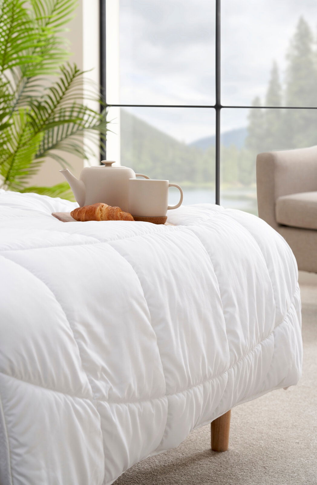 Gemütliche weiße Decke auf einem Bett mit einer Teekanne, Tasse und Croissant, im Hintergrund ein großes Fenster mit Blick auf die Natur, seitliche Perspektive.
