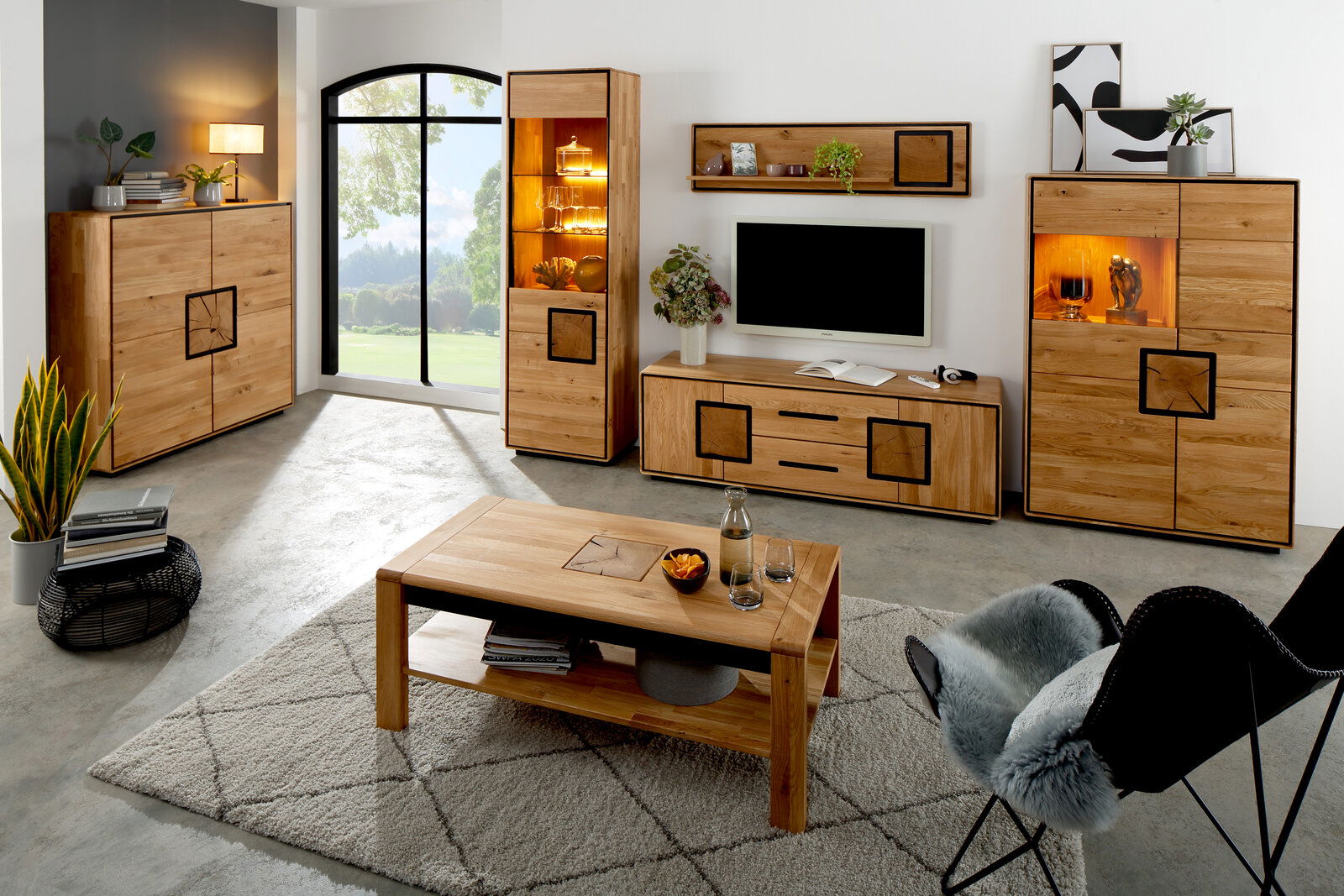 Modernes Wohnzimmer-Set aus hellem Massivholz mit Sideboard, Vitrine mit Beleuchtung, TV-Lowboard, Wandregal und Couchtisch auf Teppich, fotografiert in frontaler Raumansicht mit leichter Schrägperspektive von vorn.