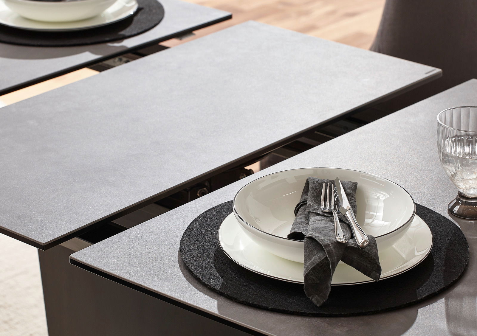 Nahaufnahme eines modernen Esstisches mit grauer Oberfläche, gedeckt mit einem Teller, Besteck und einem Glas, aus der Vogelperspektive.