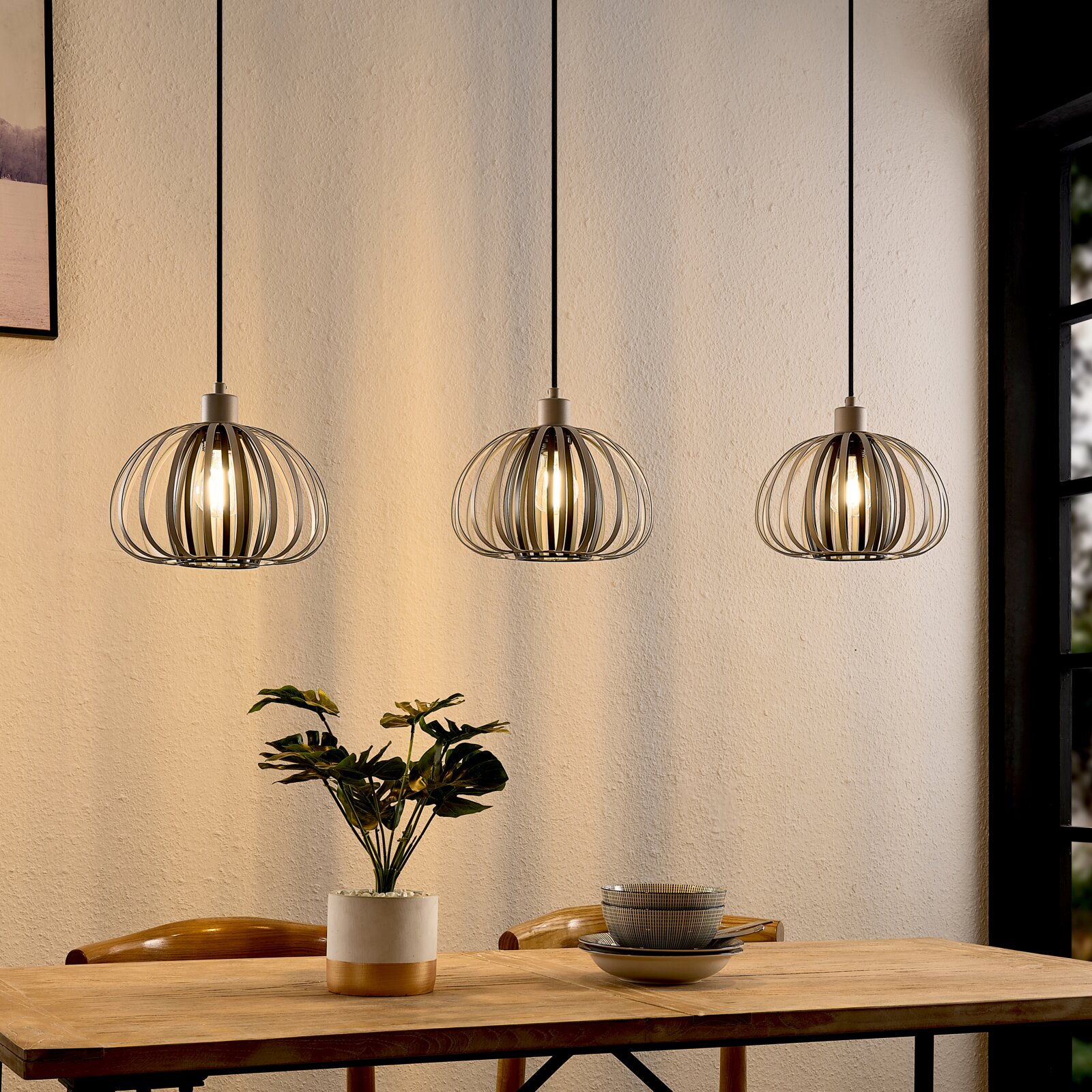 Drie moderne hanglampen met een metalen kooivormig ontwerp boven een eettafel, gezien vanaf de zijkant.