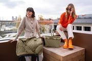Zwei Frauen auf einer Terrasse, eine sitzt auf einem Hocker neben einer olivgrünen Reisetasche, die andere sitzt auf einer Holzkiste neben einem olivgrünen Einkaufskorb. Beide Produkte haben ein Rautenmuster. Die Perspektive zeigt die Produkte von vorne.