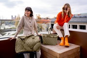 Zwei Frauen auf einer Terrasse, eine sitzt auf einem Hocker neben einer olivgrünen Reisetasche, die andere sitzt auf einer Holzkiste neben einem olivgrünen Einkaufskorb. Beide Produkte haben ein Rautenmuster. Die Perspektive zeigt die Produkte von vorne.