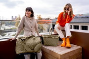 Zwei Frauen auf einer Terrasse, eine sitzt auf einem Hocker neben einer olivgrünen Reisetasche, die andere sitzt auf einer Holzkiste neben einem olivgrünen Einkaufskorb. Beide Produkte haben ein Rautenmuster. Die Perspektive zeigt die Produkte von vorne.