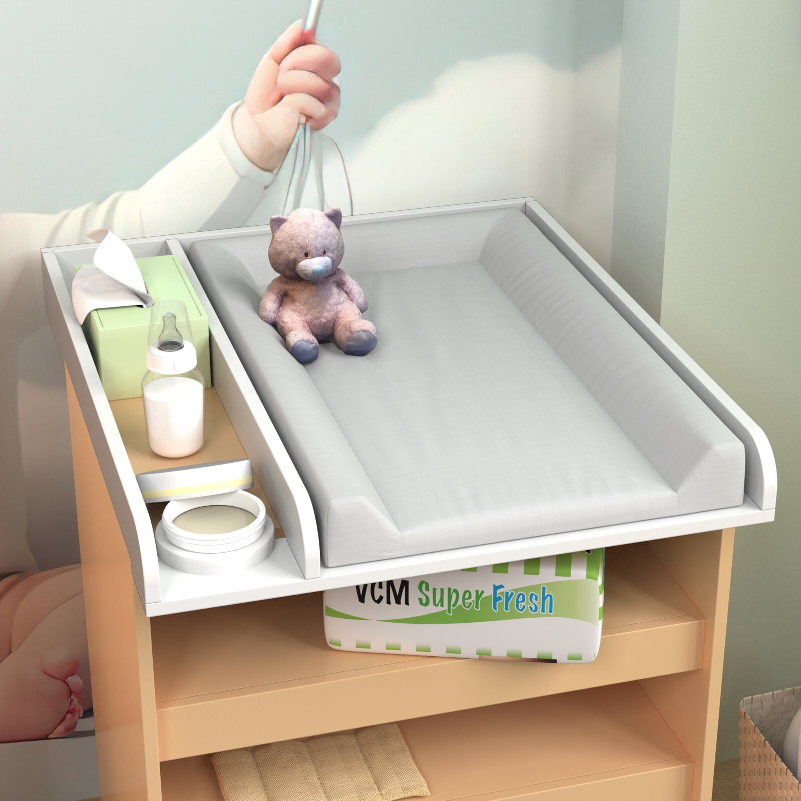 Wickelaufsatz für eine Kommode mit gepolsterter grauer Wickelauflage, seitlichem Ablagefach mit Feuchttüchern und Babyflasche sowie Teddy; Ansicht leicht von oben in schräger Perspektive.