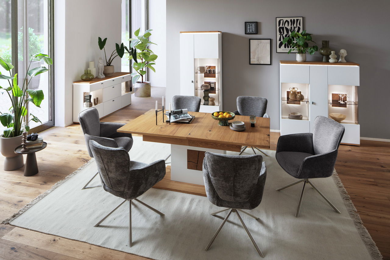 Modernes Esszimmer mit einem großen Holztisch, umgeben von grauen Stühlen. Im Hintergrund sind ein Sideboard und zwei Vitrinen mit Dekorationen zu sehen. Die Perspektive zeigt den Raum von der Seite mit Blick auf den Tisch und die Möbel.