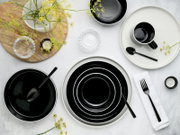 Schwarz-weißes Keramik-Geschirrset mit Tellern, Schalen und Tasse sowie schwarzem Besteck auf heller Tischfläche, Draufsicht (von oben)