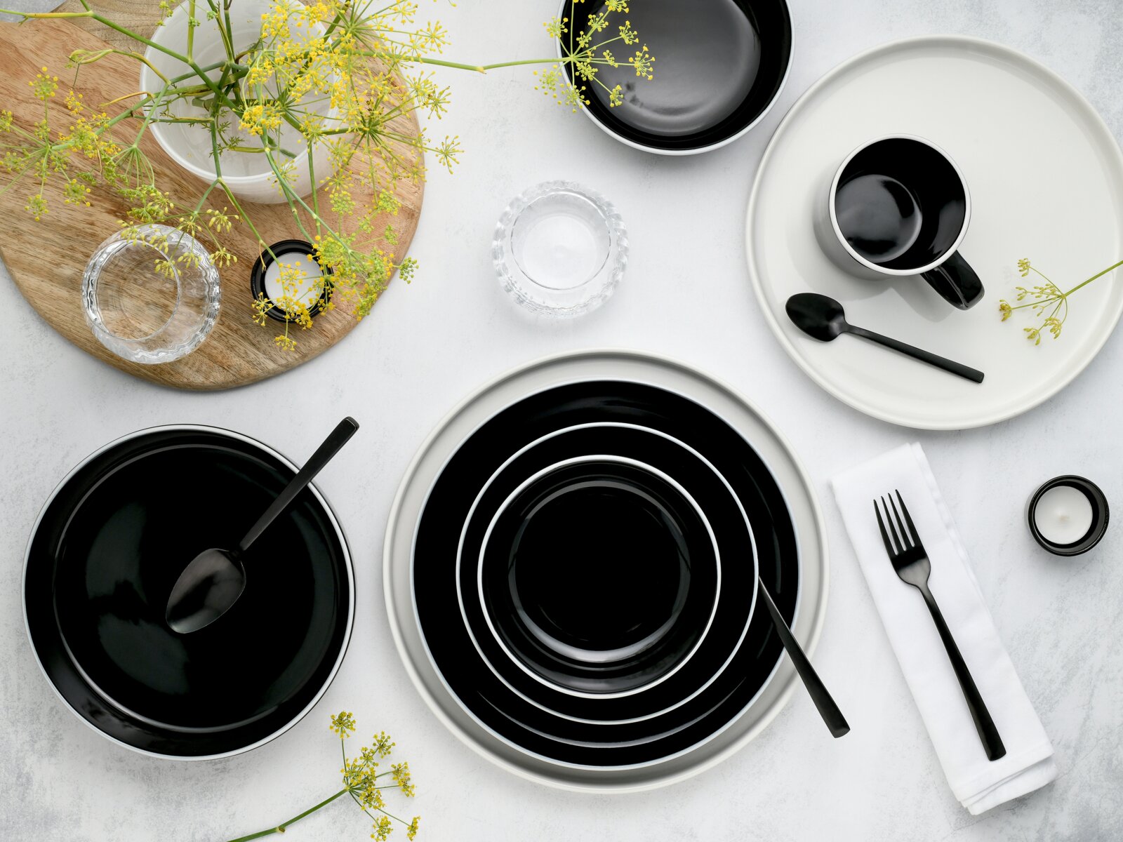 Schwarz-weißes Keramik-Geschirrset mit Tellern, Schalen und Tasse sowie schwarzem Besteck auf heller Tischfläche, Draufsicht (von oben)