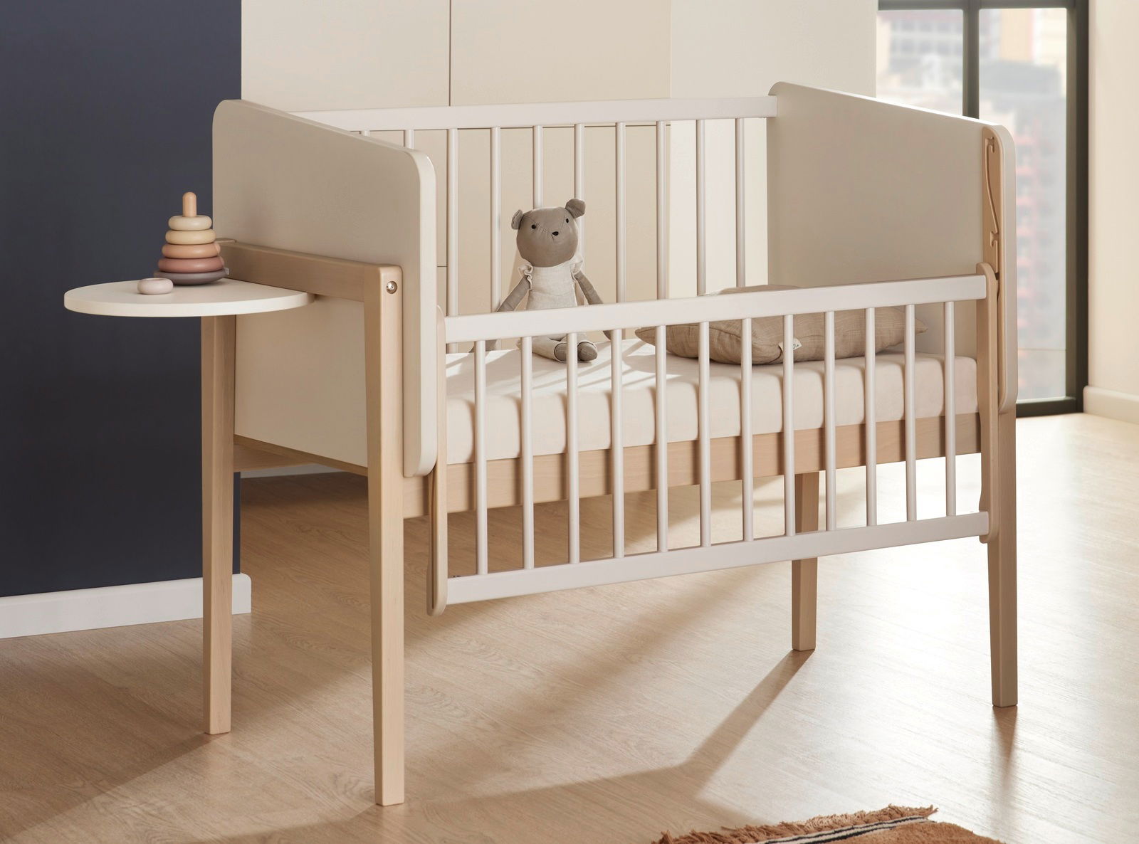 Kinderbett mit seitlichem Tablar, von schräg vorne fotografiert, mit einem Stofftier und einem Stapelspielzeug.