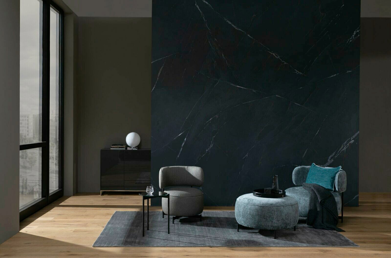 Moderner Wohnbereich mit einem grauen Sessel, einem runden Hocker und einem kleinen runden Beistelltisch auf einem Teppich vor einer dunklen Wand. Perspektive von vorne.