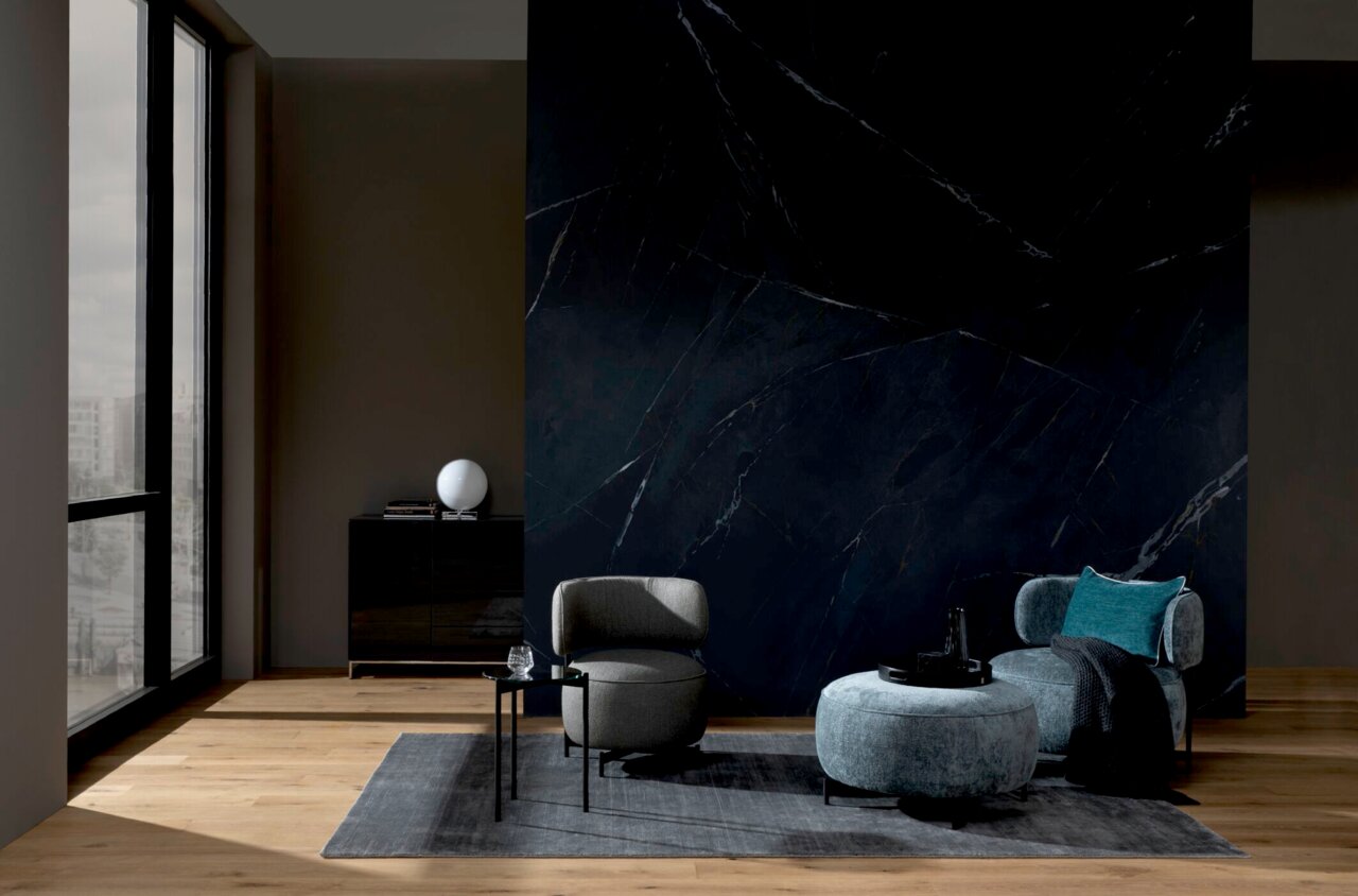 Moderner Wohnbereich mit einem grauen Sessel, einem runden Hocker und einem kleinen runden Beistelltisch auf einem Teppich vor einer dunklen Wand. Perspektive von vorne.