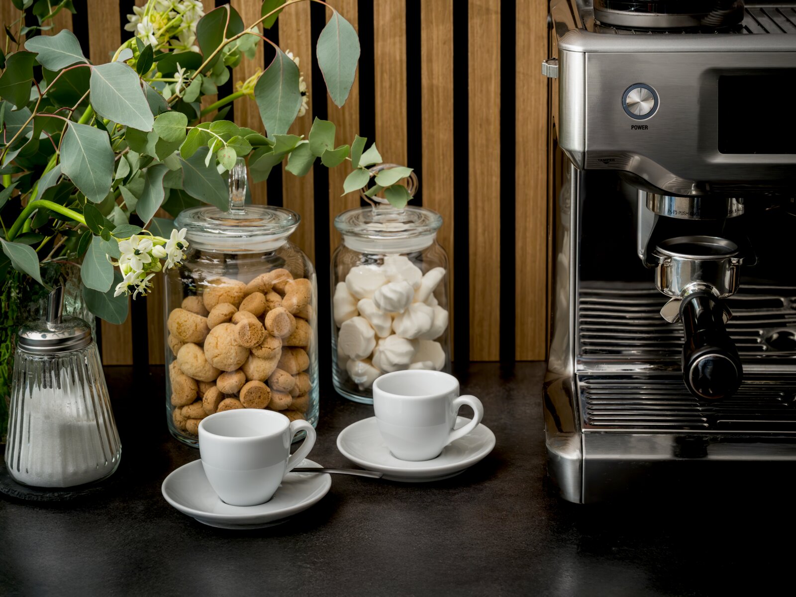 Creatable Espresso Set 8-tlg BISTROT Küchen- bzw. Kaffeestation in frontaler Perspektive: Edelstahl-Espressomaschine rechts, davor zwei weiße Espressotassen mit Untertassen; daneben Glasbehälter mit Keksen und Marshmallows sowie grüne Zweige und Zuckerdose auf dunkler Arbeitsplatte.