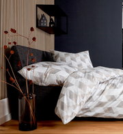 Bettwäsche-Set in geometrischem Muster auf einem grauen Bett, seitliche Perspektive, mit dekorativem Regal und Trockenblumen im Hintergrund.