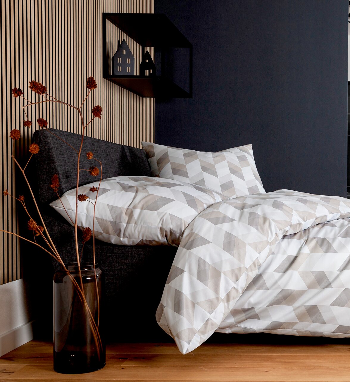 Bettwäsche-Set in geometrischem Muster auf einem grauen Bett, seitliche Perspektive, mit dekorativem Regal und Trockenblumen im Hintergrund.