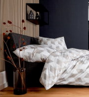 Bettwäsche-Set in geometrischem Muster auf einem grauen Bett, seitliche Perspektive, mit dekorativem Regal und Trockenblumen im Hintergrund.