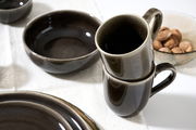 Nahaufnahme von gestapelten, dunkelgrauen Bechern auf einem Tisch, umgeben von einer Schale und einem Teller, mit einer Schale voller Nüsse im Hintergrund.