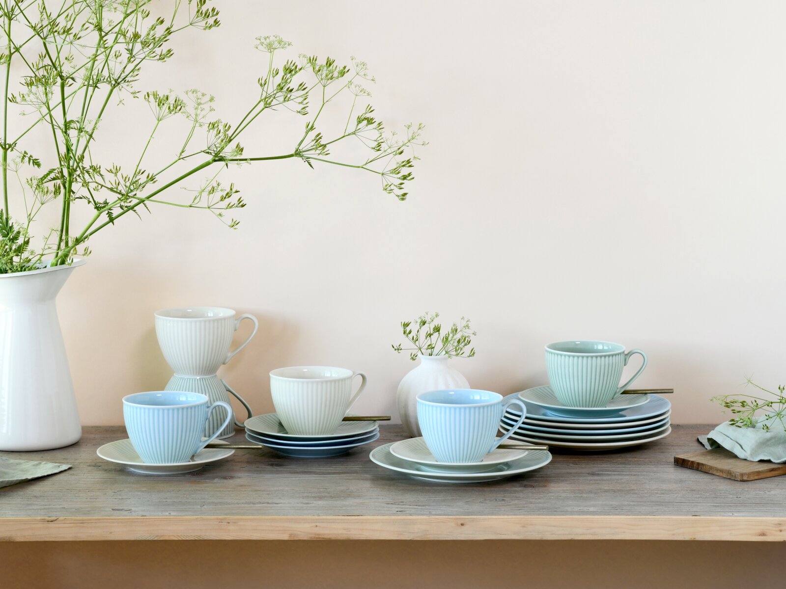 Pastellfarbenes Kaffee- und Tafelservice mit geriffelten Tassen und passenden Untertassen auf Holztisch, seitliche Perspektive auf die gedeckte Szene vor hellem Hintergrund