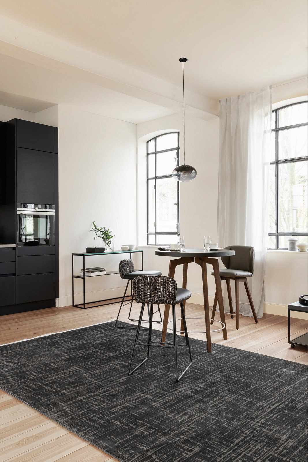 Moderner Essbereich mit einem schwarzen Teppich von JOOP! in einem hellen Raum mit hohen Fenstern, stilvollen Barhockern und einem runden Tisch, aus der Perspektive des Raumes aufgenommen.