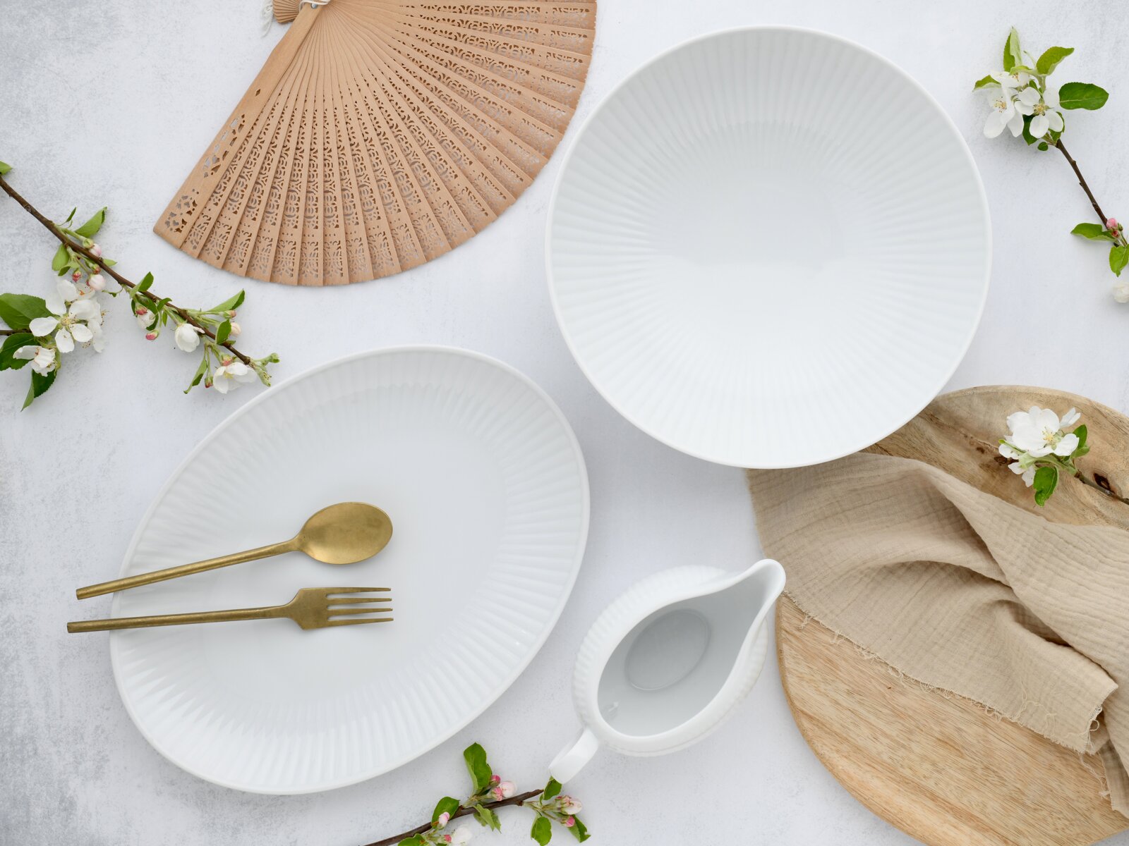 Draufsicht auf ein minimalistisches Tisch-Arrangement mit zwei weißen, geriffelten Porzellanschalen, weißem Milchkännchen, goldfarbenem Löffel und Gabel, Holzbrett, beigefarbenem Tuch, Papierfächer und Frühlingsblüten auf hellem Hintergrund