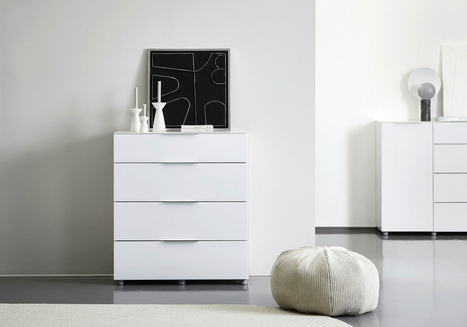 Voleo Schubkastenkommode MALIN Weiße Schubkastenkommode mit vier Schubladen, frontal fotografiert, dekoriert mit modernen Kerzenhaltern und einem abstrakten Kunstwerk.