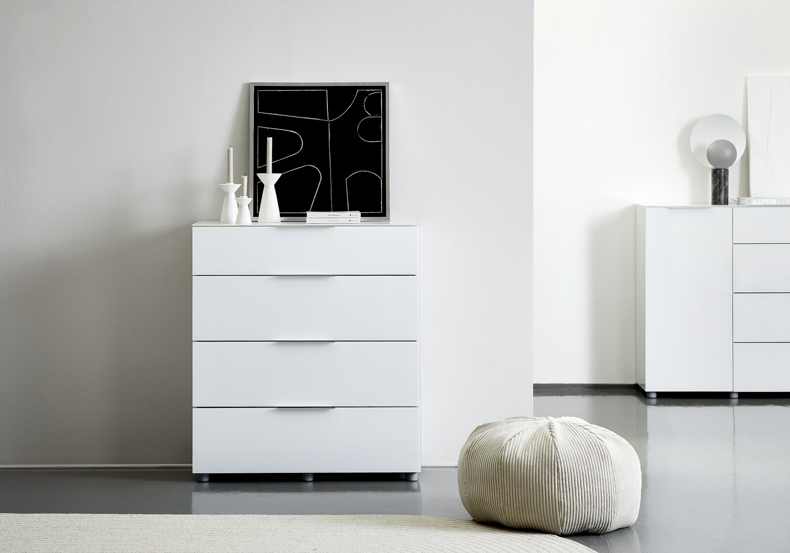 Voleo Schubkastenkommode MALIN Weiße Schubkastenkommode mit vier Schubladen, frontal fotografiert, dekoriert mit modernen Kerzenhaltern und einem abstrakten Kunstwerk.