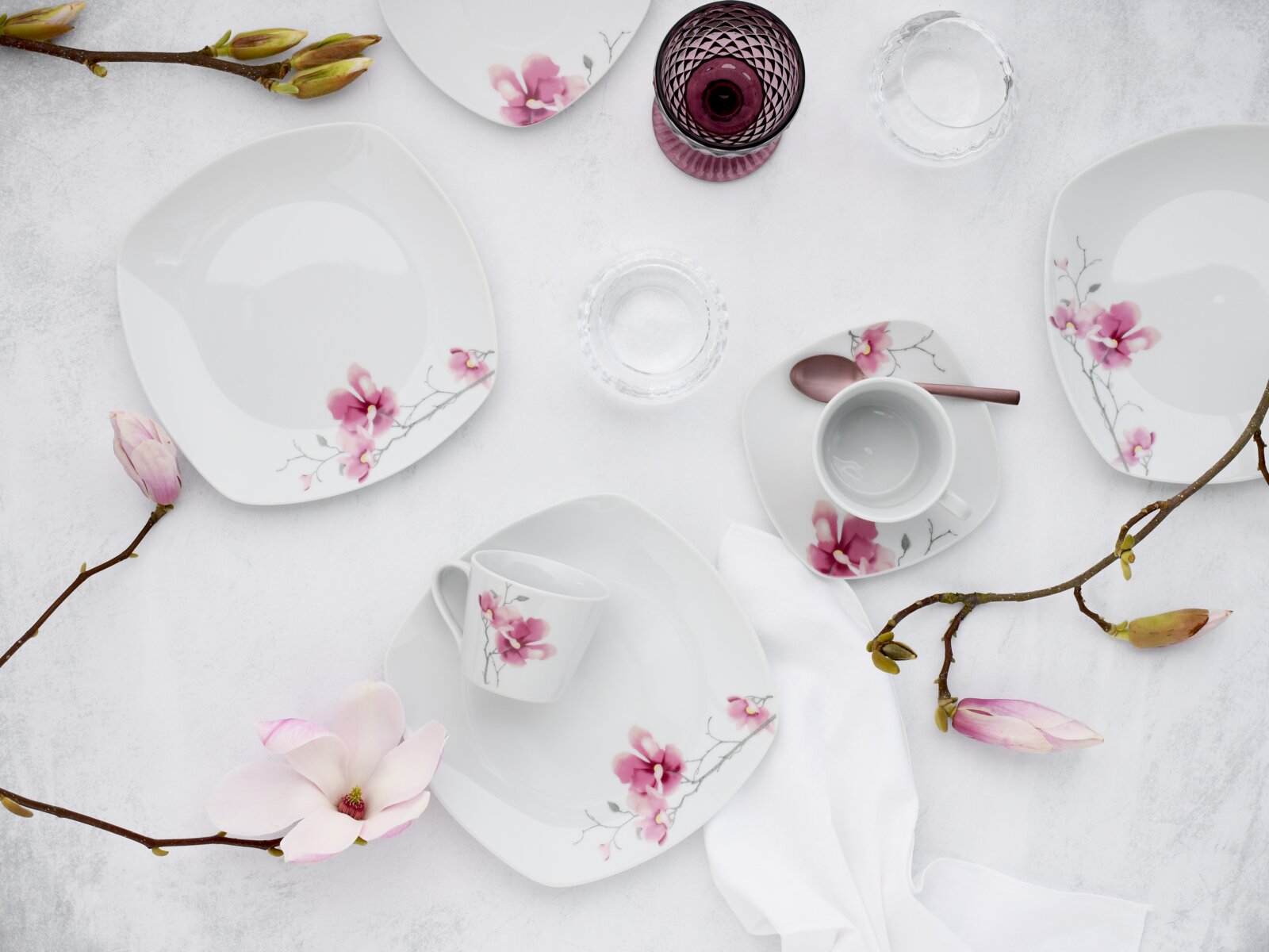 Porzellan-Geschirrset in Weiß mit rosa Blütenmotiv (Teller, Schalen und Tasse) dekorativ arrangiert auf hellem Untergrund mit Magnolienzweigen, Ansicht von oben (Flatlay).