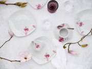 Porzellan-Geschirrset in Weiß mit rosa Blütenmotiv (Teller, Schalen und Tasse) dekorativ arrangiert auf hellem Untergrund mit Magnolienzweigen, Ansicht von oben (Flatlay).