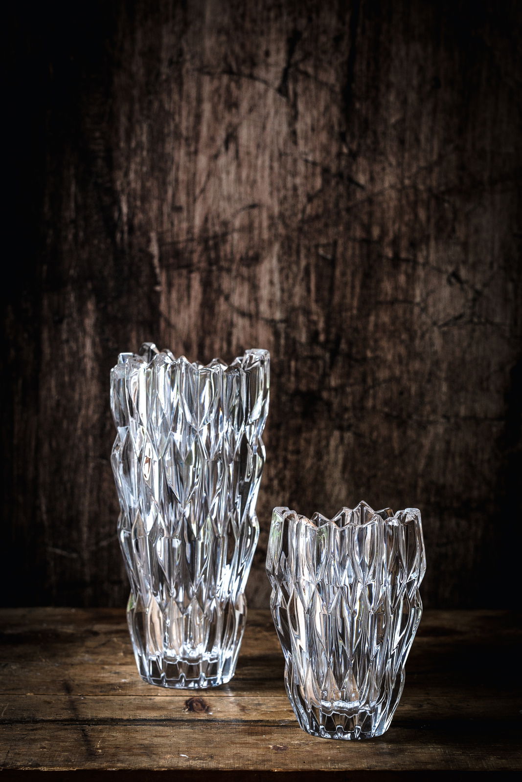 Zwei kristallklare Vasen mit facettierter Oberfläche, eine größere und eine kleinere, auf einem rustikalen Holztisch vor einem dunklen Hintergrund. Perspektive von vorne.