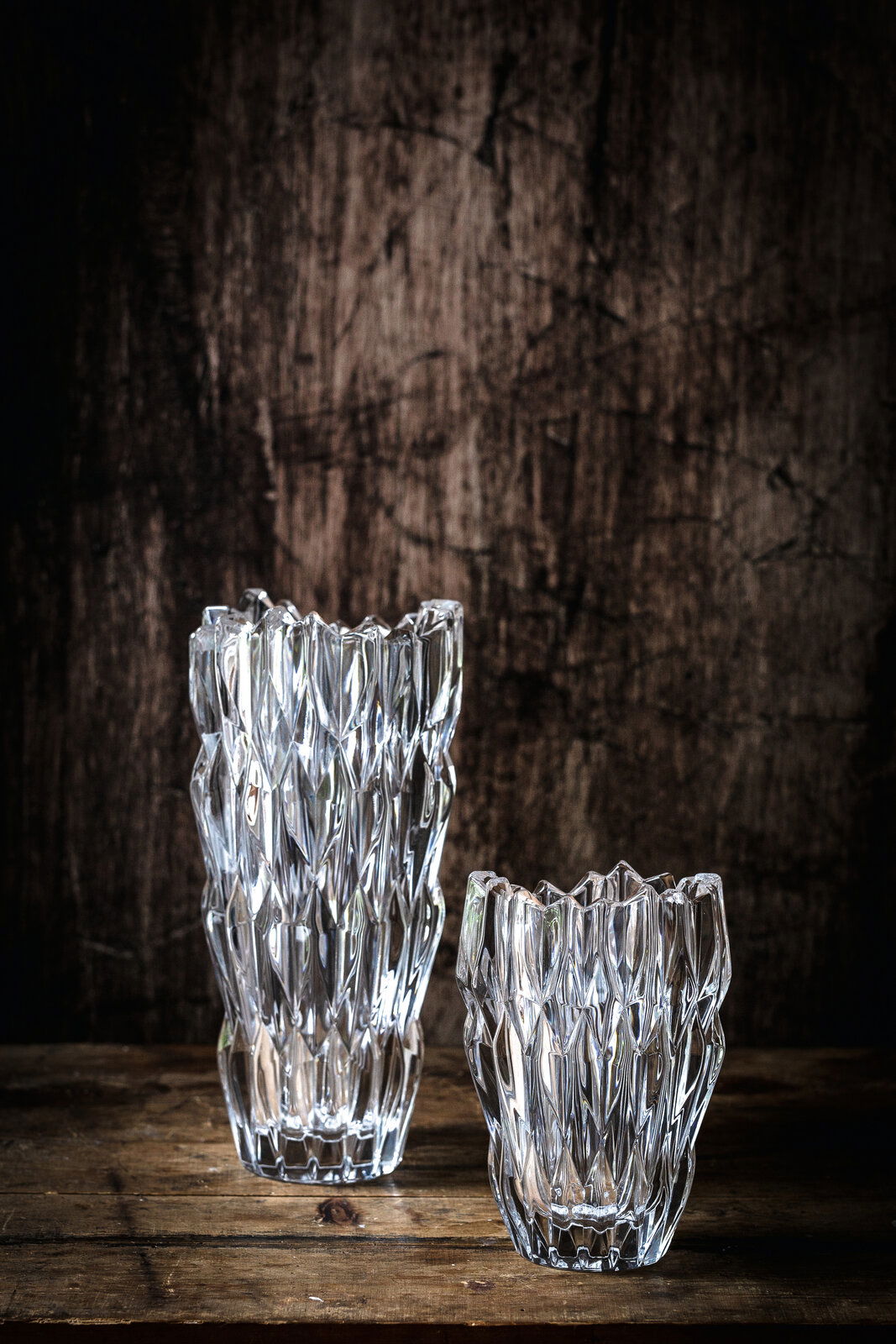 Zwei kristallklare Vasen mit facettierter Oberfläche, eine größere und eine kleinere, auf einem rustikalen Holztisch vor einem dunklen Hintergrund. Perspektive von vorne.