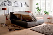 Polsterbett mit hochgeklappter Matratze, seitliche Perspektive, zeigt Stauraum darunter, umgeben von stilvoller Dekoration und Pflanzen.