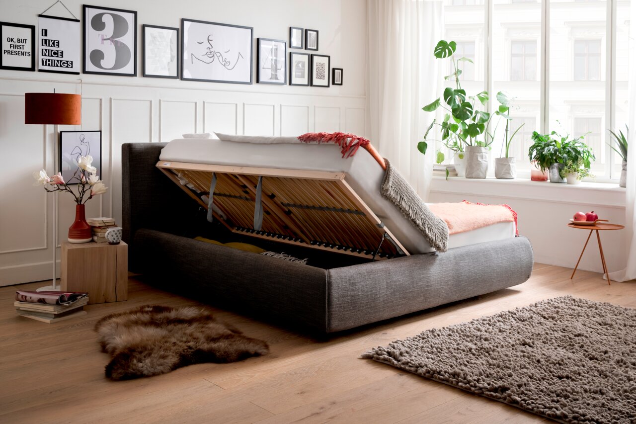 Polsterbett mit hochgeklappter Matratze, seitliche Perspektive, zeigt Stauraum darunter, umgeben von stilvoller Dekoration und Pflanzen.