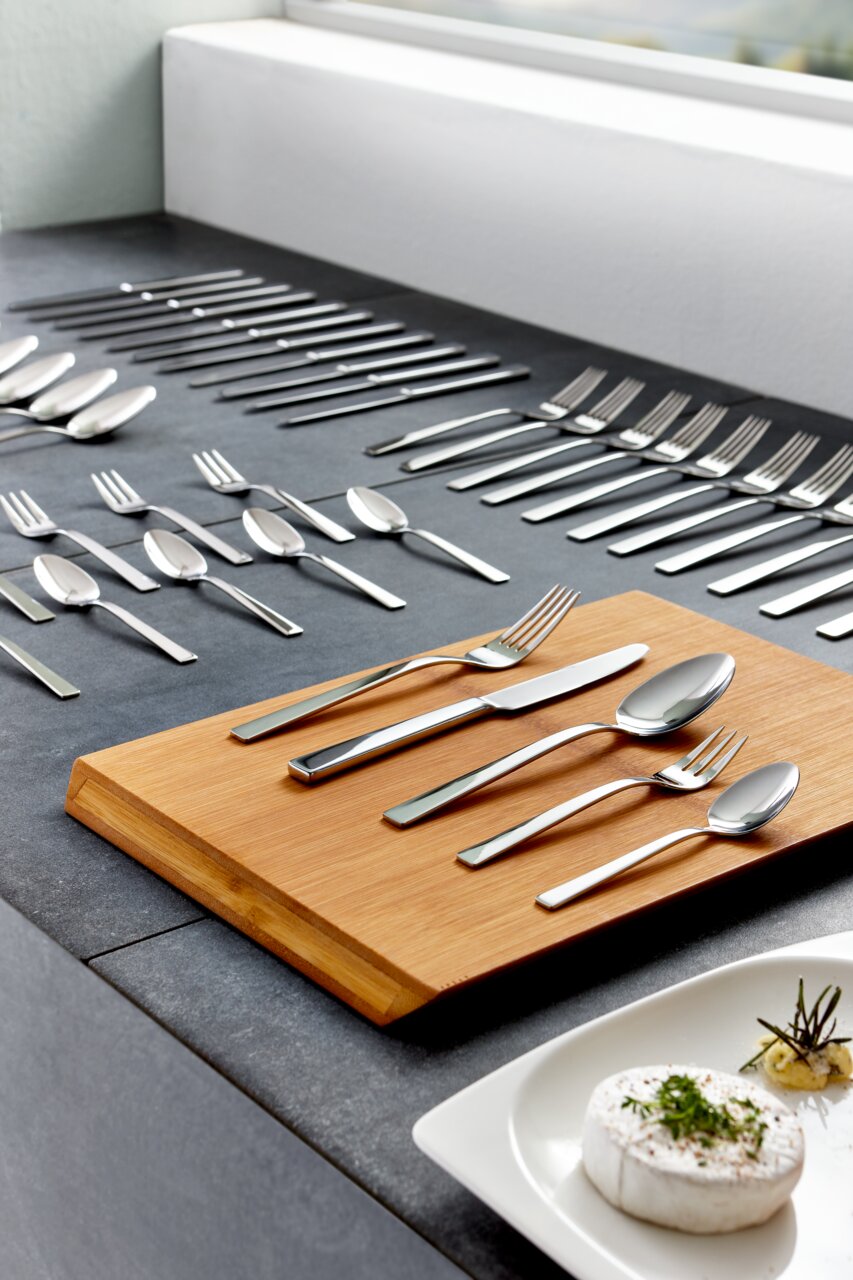 Elegantes 60-teiliges poliertes Besteckset 'King' auf einem Tisch, mit einem Holzbrett im Vordergrund und einer modernen, stilvollen Anordnung. Perspektive von schräg oben.