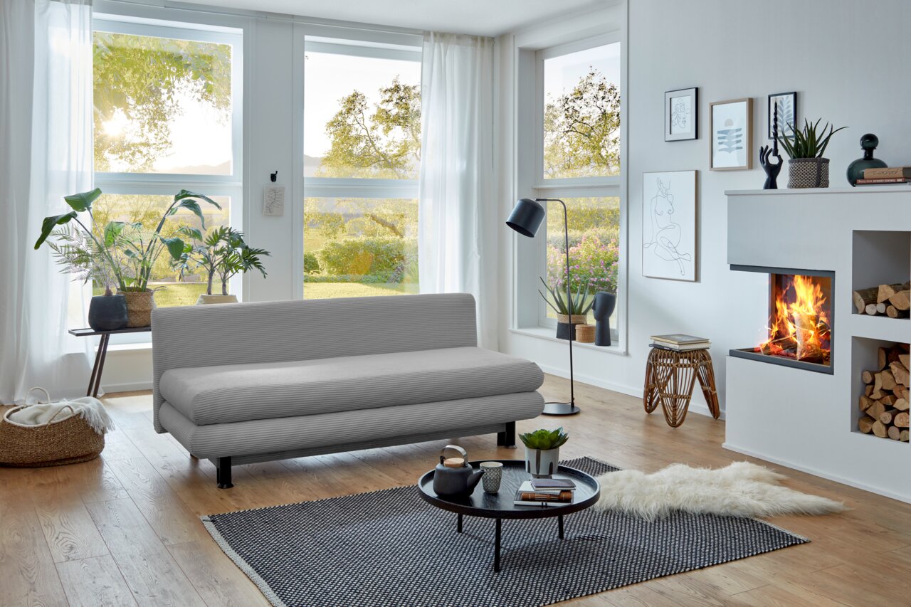Graues Funktionssofa in modernem Wohnzimmer mit großen Fenstern, Pflanzen und Kamin, seitliche Perspektive.
