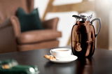 Isolierkanne Juwel in glänzendem Braun auf einem Tisch, mit einer Tasse Kaffee und einem Keks im Vordergrund, seitliche Perspektive.