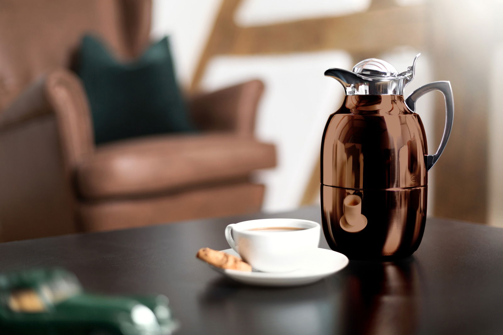 Isolierkanne Juwel in glänzendem Braun auf einem Tisch, mit einer Tasse Kaffee und einem Keks im Vordergrund, seitliche Perspektive.