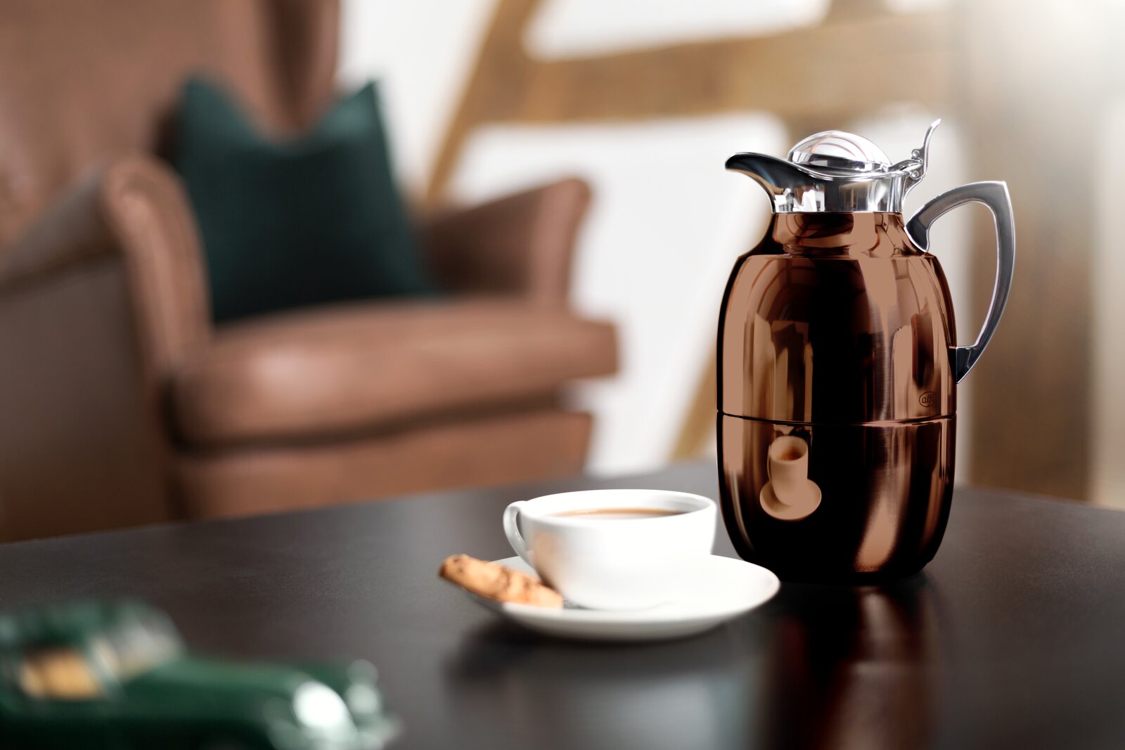 Isolierkanne Juwel in glänzendem Braun auf einem Tisch, mit einer Tasse Kaffee und einem Keks im Vordergrund, seitliche Perspektive.