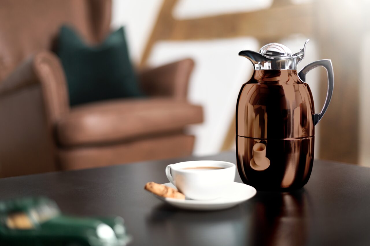 Isolierkanne Juwel in glänzendem Braun auf einem Tisch, mit einer Tasse Kaffee und einem Keks im Vordergrund, seitliche Perspektive.