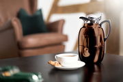 Isolierkanne Juwel in glänzendem Braun auf einem Tisch, mit einer Tasse Kaffee und einem Keks im Vordergrund, seitliche Perspektive.