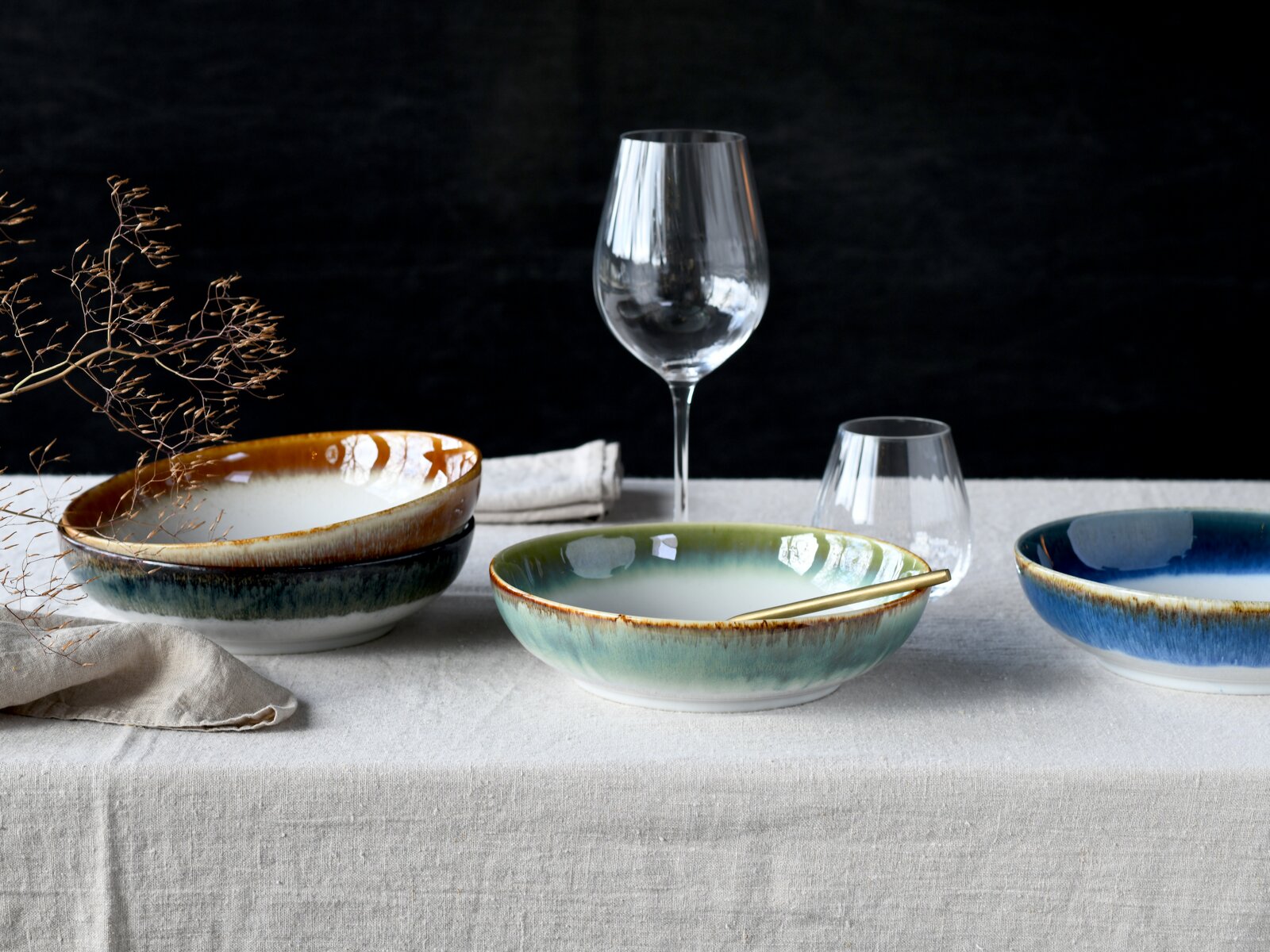 Modernes Tisch-Arrangement mit drei glasierten Keramikschalen in Grün-, Blau- und Braunverlauf auf Leinentischdecke, dazu Weinglas, Wasserglas und goldfarbener Löffel; Perspektive: frontale Ansicht auf Augenhöhe mit leichtem Abstand.