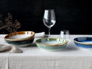 Modernes Tisch-Arrangement mit drei glasierten Keramikschalen in Grün-, Blau- und Braunverlauf auf Leinentischdecke, dazu Weinglas, Wasserglas und goldfarbener Löffel; Perspektive: frontale Ansicht auf Augenhöhe mit leichtem Abstand.