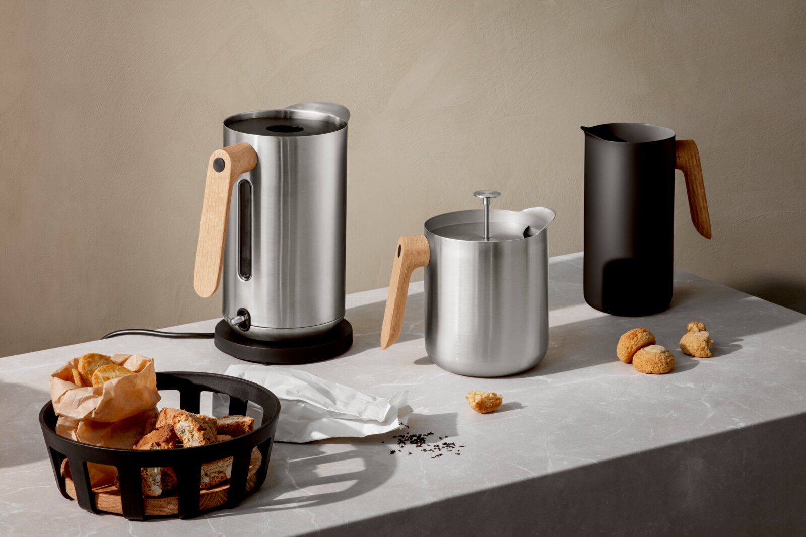 Eva Solo Wasserkocher NORDIC KITCHEN Schräg von vorn fotografiertes Set aus Edelstahl-Wasserkocher mit Holzgriff, Edelstahl-Milchaufschäumer mit Stempel und schwarzem Milchkrug mit Holzgriff auf heller Küchenarbeitsplatte, daneben Gebäck und Brot im Korb.