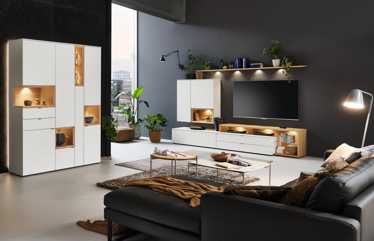 Moderne Wohnzimmeransicht mit der Vitrine Venjakob ANDIAMO HOME in Weiß, beleuchteten Fächern und stilvoller Dekoration, seitliche Perspektive.