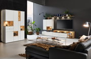 Een moderne woonkamer met de vitrinekast Venjakob ANDIAMO HOME in wit, verlichte vakken en stijlvolle decoratie, gezien vanuit de zijkant.
