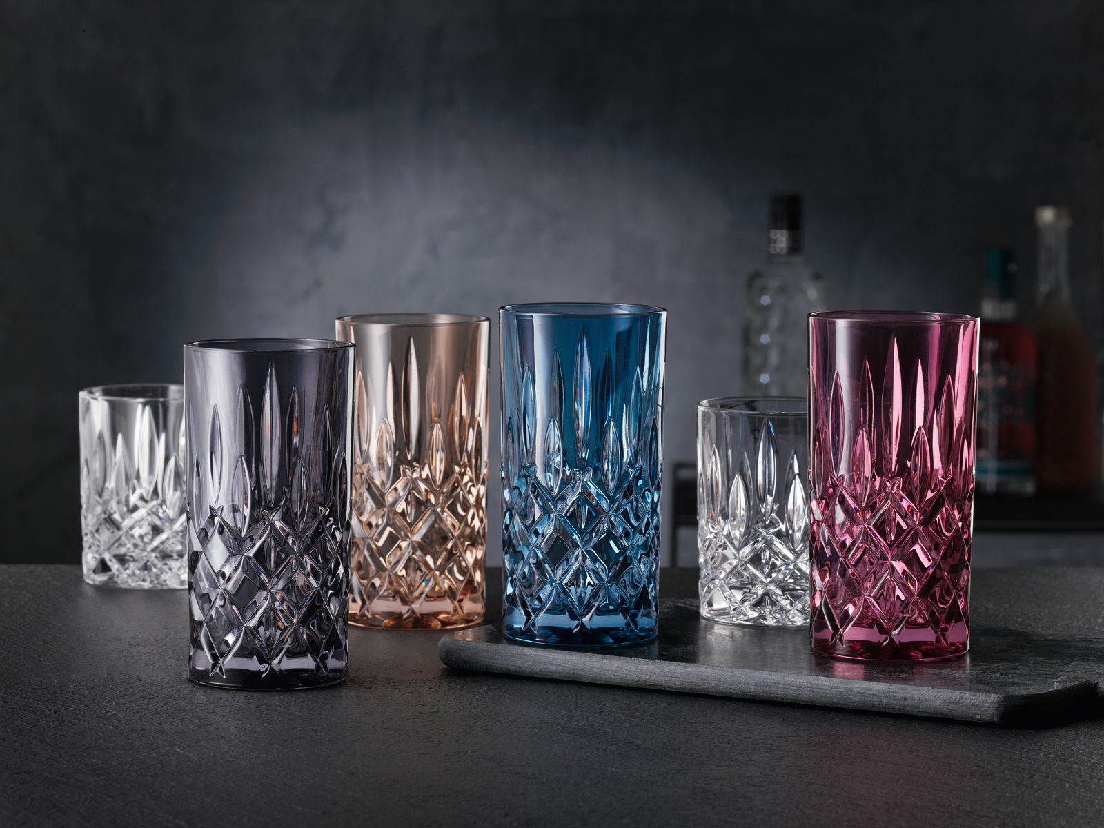 Verschiedene farbige Longdrinkgläser aus Kristallglas in edlem Design, frontal auf einem dunklen Tisch arrangiert.