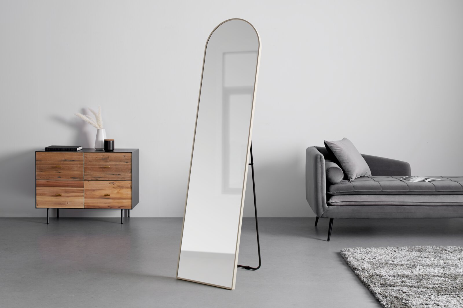 Schlanker, freistehender Standspiegel mit abgerundeter Oberkante und hellem Rahmen, leicht schräg von vorne in einem minimalistischen Wohnzimmer gezeigt