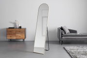 Schlanker, freistehender Standspiegel mit abgerundeter Oberkante und hellem Rahmen, leicht schräg von vorne in einem minimalistischen Wohnzimmer gezeigt