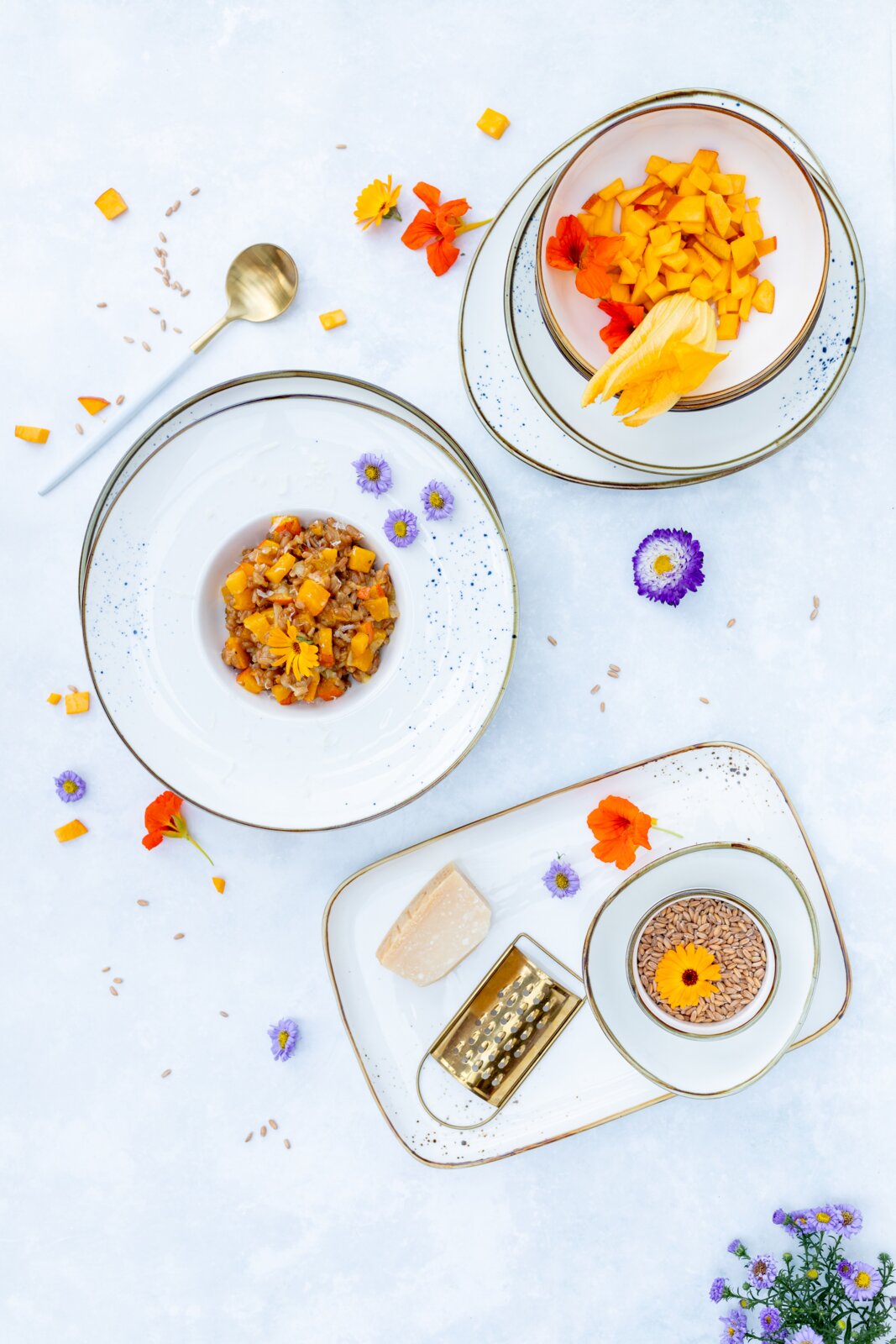Draufsicht auf ein elegantes weißes Geschirr-Arrangement mit gesprenkeltem Rand: Schale mit gewürfelter Mango und essbaren Blüten, Teller mit herzhaftem Reis-Gemüse-Mix, dazu Löffel, Parmesan, Reibe und Schälchen mit Körnern auf hellem Hintergrund.