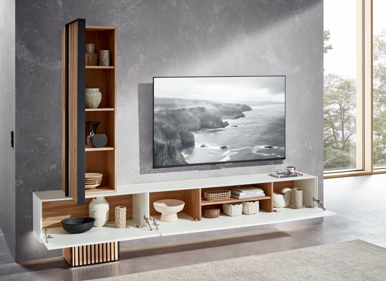 Moderne wandcombinatie met open planken en gesloten vakken, versierd met keramiek en manden, naast een grote tv tegen een grijze muur. Aanzicht van schuin van voren.