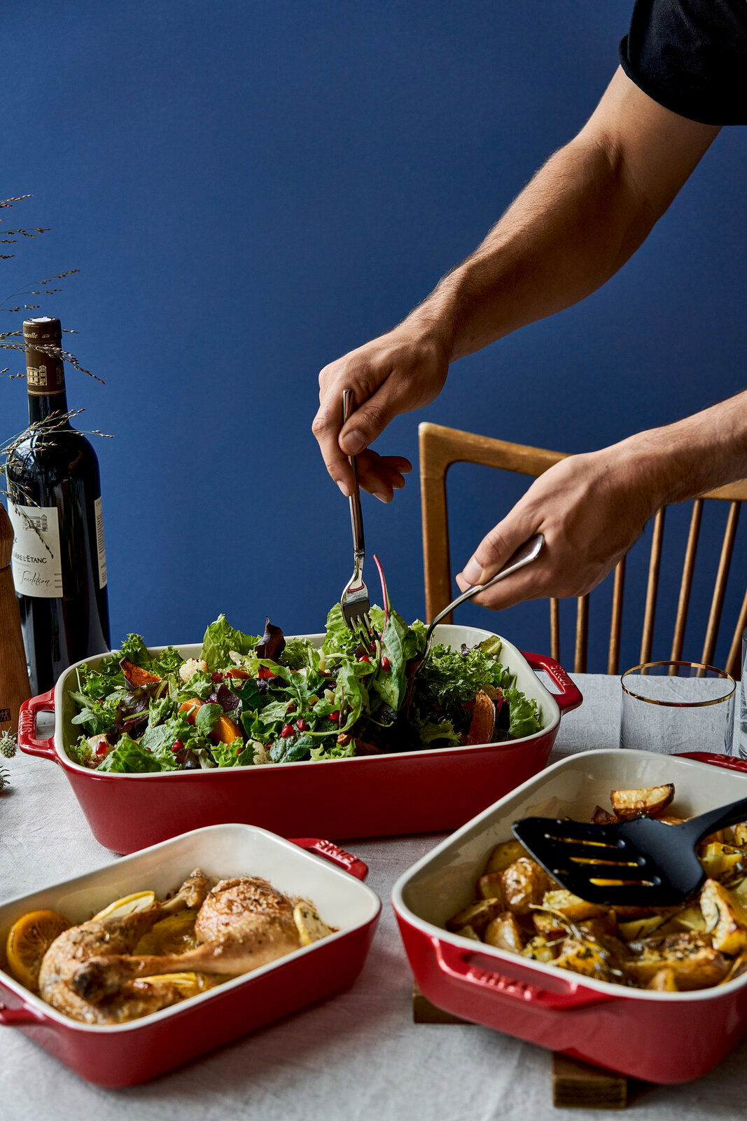 Drei rote Auflaufformen mit verschiedenen Gerichten, darunter ein frischer Salat, Hähnchenschenkel und Ofenkartoffeln, auf einem Tisch von oben fotografiert.