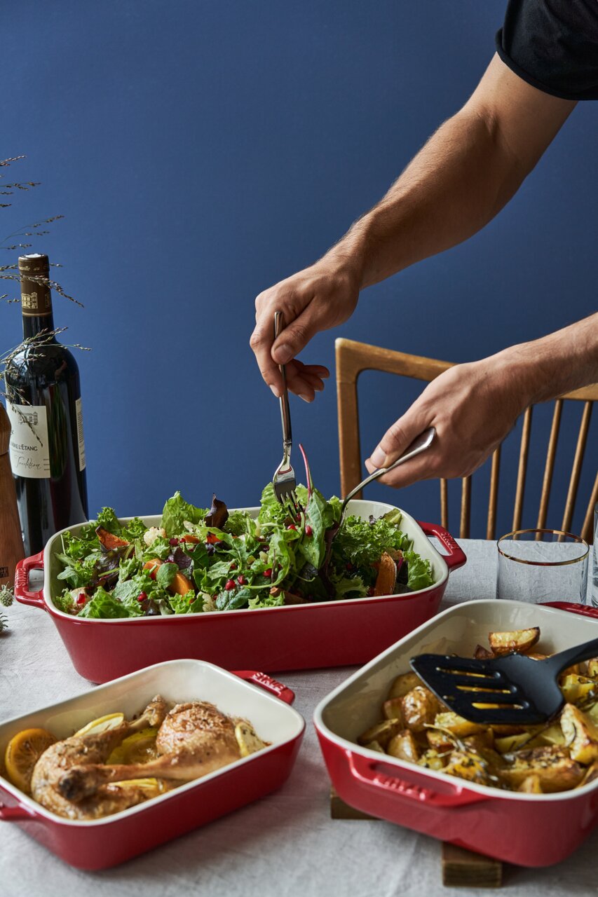 Drie rode ovenschaaltjes met verschillende gerechten, waaronder een frisse salade, kippenboutjes en gepofte aardappelen, van bovenaf gefotografeerd op een tafel.